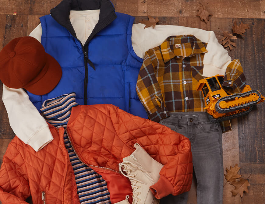 A collection of autumn clothing and accessories displayed on a wooden floor, including a blue vest, a plaid shirt, a red quilted jacket, grey jeans, a brown hat, white boots, brown shoes, and a toy mini bulldozer, all surrounded by scattered leaves