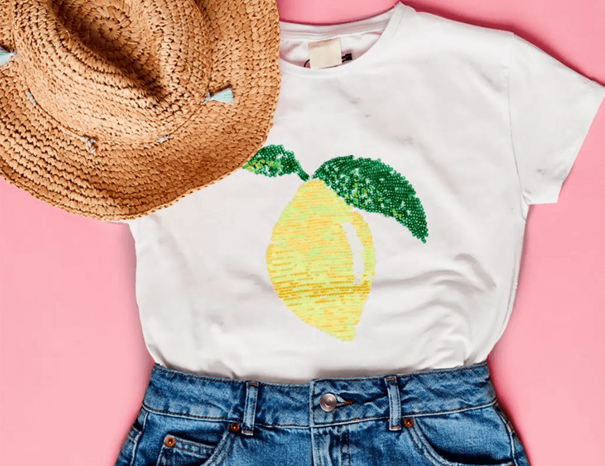 Flat lay of a casual outfit comprising a t-shirt with a sequined lemon design, denim shorts, and a straw hat, arranged on a pink background.