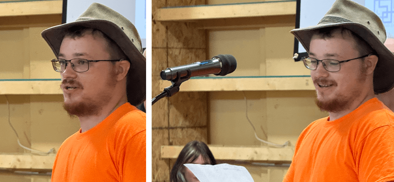 Person in an orange shirt and a hat speaking at a microphone with shelving in the background.