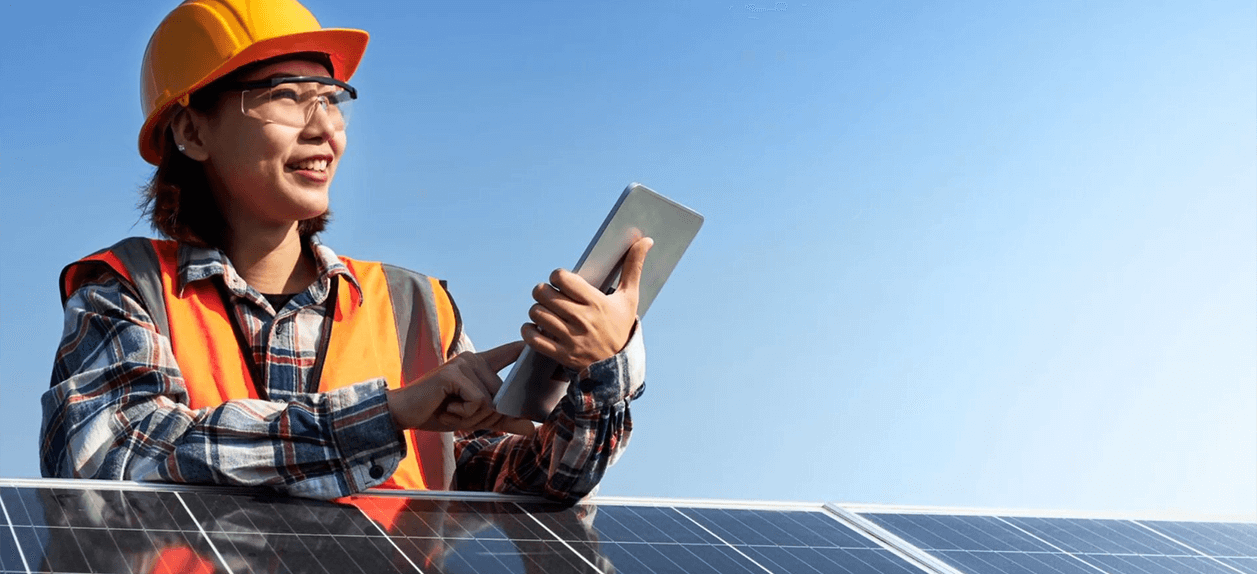 A woman stands over a solar panel wearing a hard hat and safety vest, holding a tablet