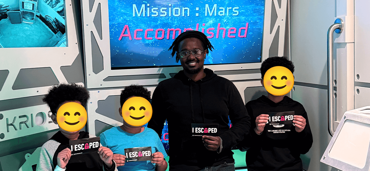 Four individuals are smiling and holding signs saying "I ESCAPED" in front of a screen displaying the message "Mission: Mars Accomplished." They are standing in a room with a futuristic, space-themed decor.